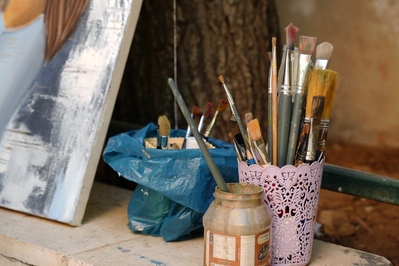 A collection of paint brushes beside an unfinished canvas outdoors in Damascus, Syria.